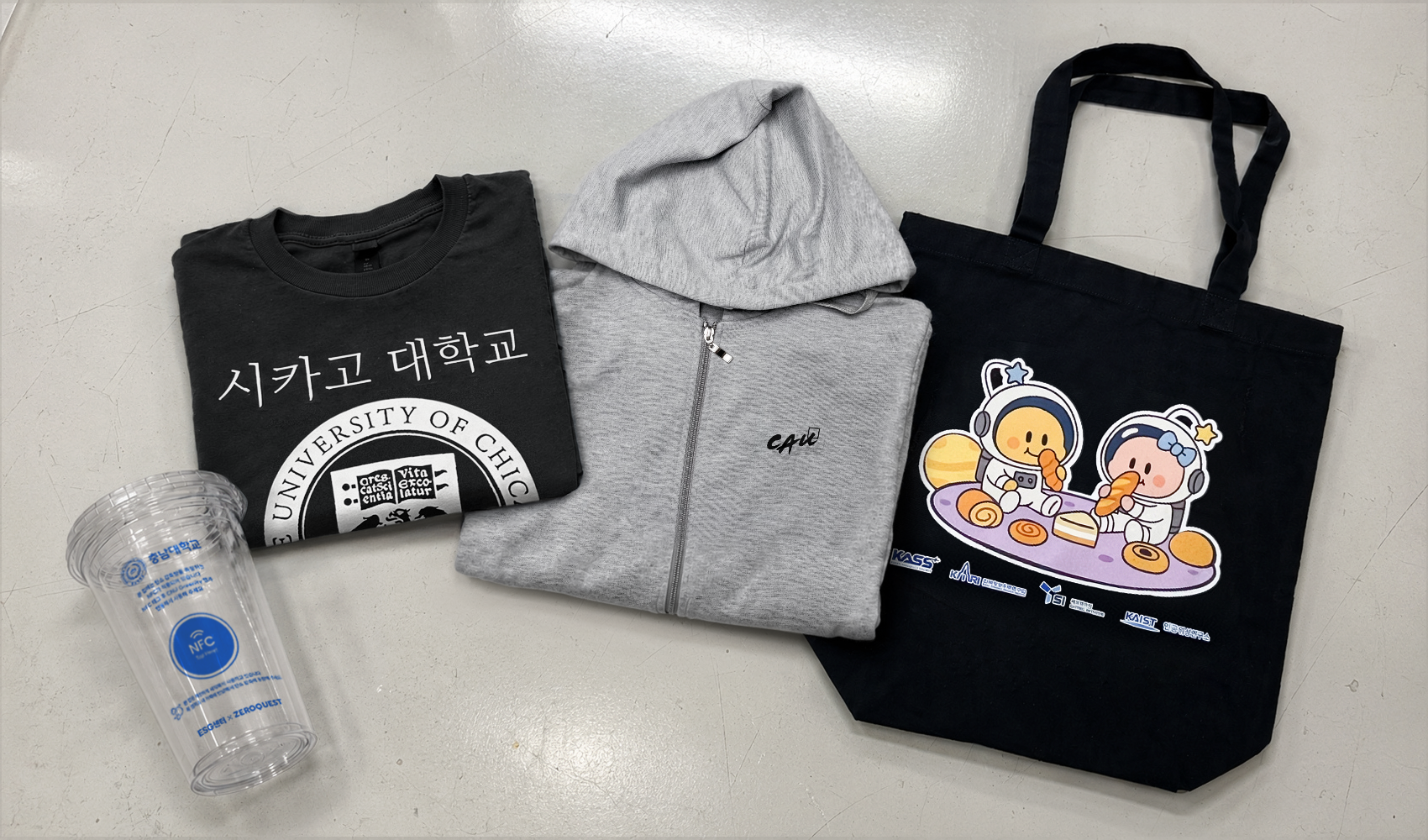 마플 대학교 단체 굿즈 제작 사례 – 시카고대학교와 KAIST 로고를 인쇄한 반팔 티셔츠, 충남대학교 NFC 리유저블컵, 우주 캐릭터 에코백을 회색 바닥에 함께 배치한 이미지
