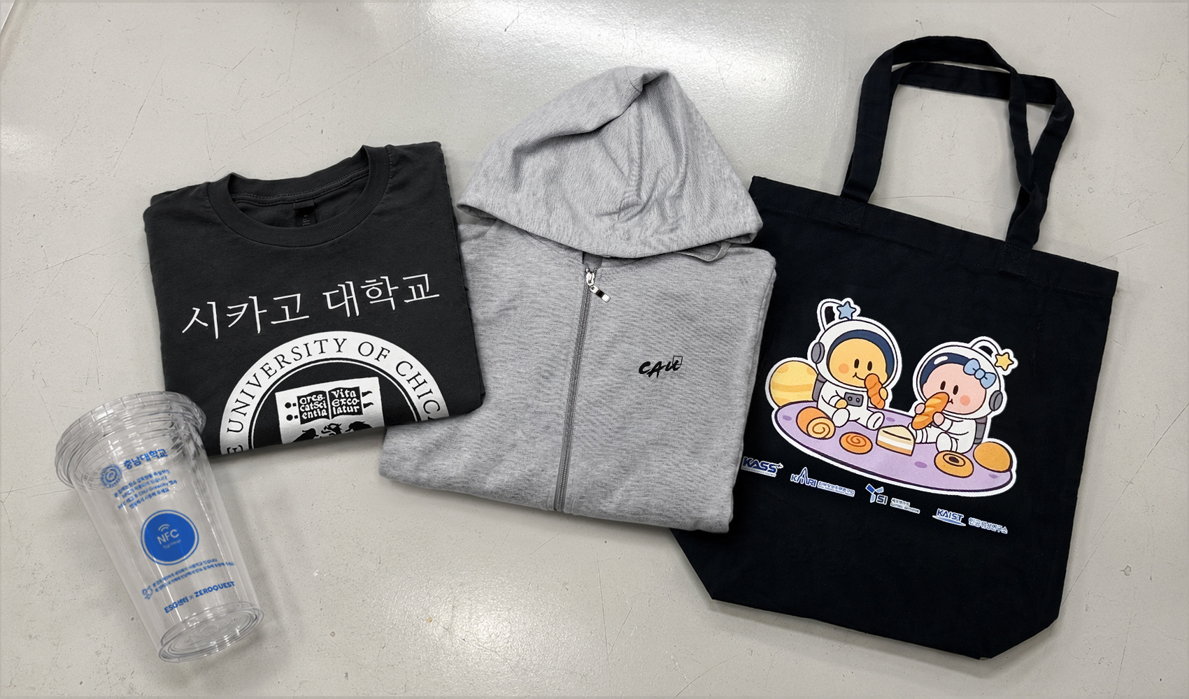 마플 대학교 단체 굿즈 제작 사례 – 시카고대학교와 KAIST 로고를 인쇄한 반팔 티셔츠, 충남대학교 NFC 리유저블컵, 우주 캐릭터 에코백을 회색 바닥에 함께 배치한 이미지