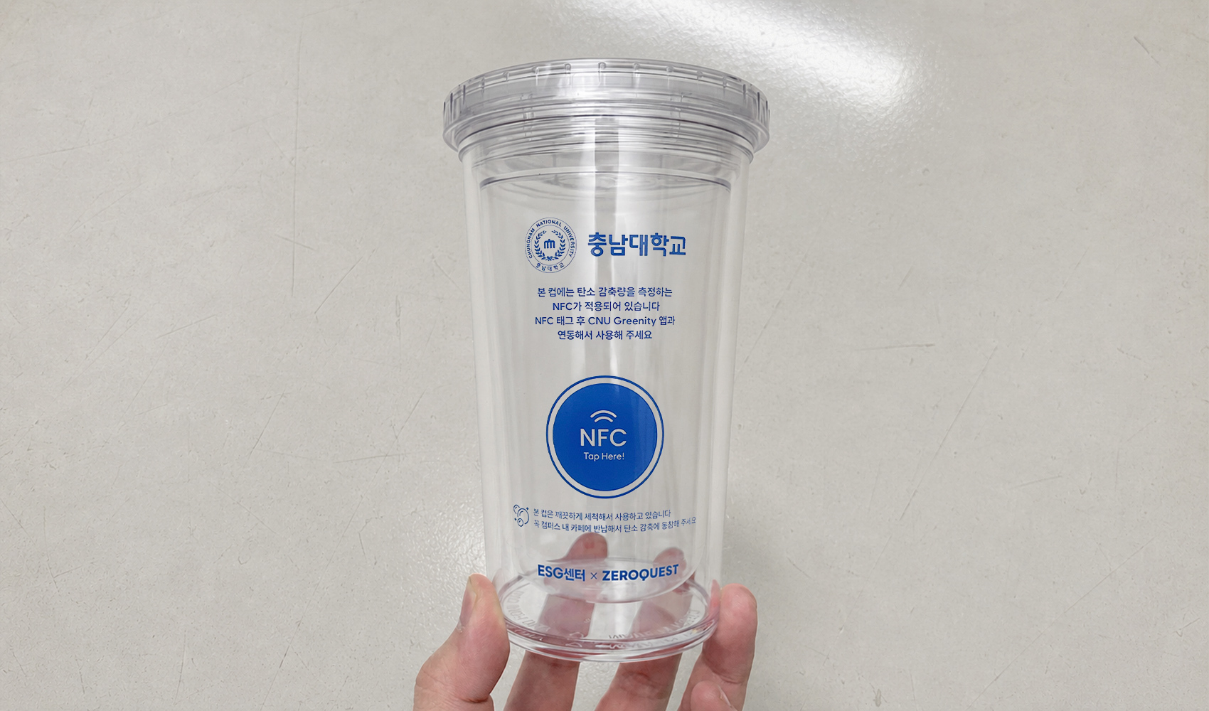 마플 NFC 리유저블컵 – 투명 컵 전면에 충남대학교 로고와 NFC 태그 안내 문구를 파란색으로 인쇄해 회색 바닥에 놓은 이미지