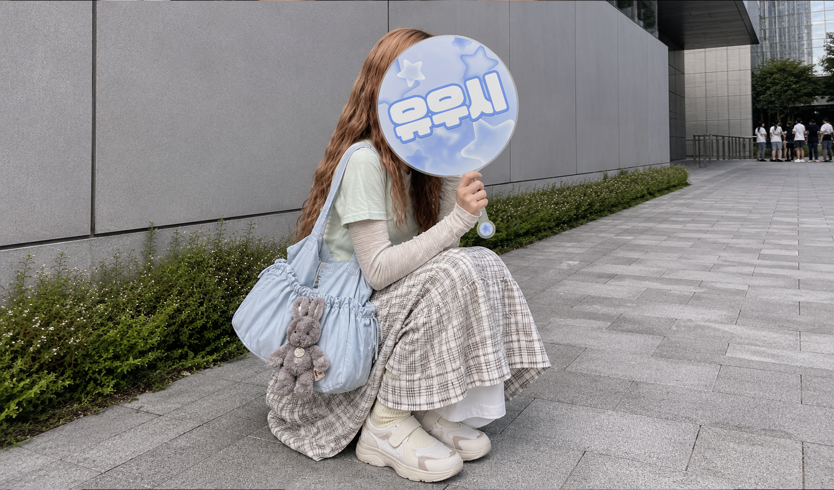 마플 대형 이미지 피켓 – 최애 이름과 별 그래픽을 하늘색으로 인쇄한 원형 우치와로 얼굴을 가리고 콘서트장 앞에서 포즈를 취한 여성 이미지
