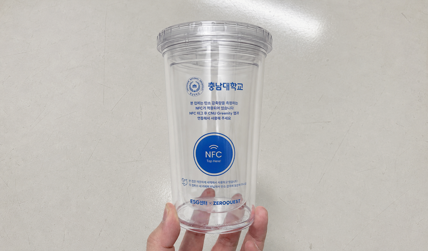 마플 NFC 리유저블컵 – 투명 컵 전면에 충남대학교 로고와 NFC 태그 안내 문구를 파란색으로 인쇄해 회색 바닥에 놓은 이미지