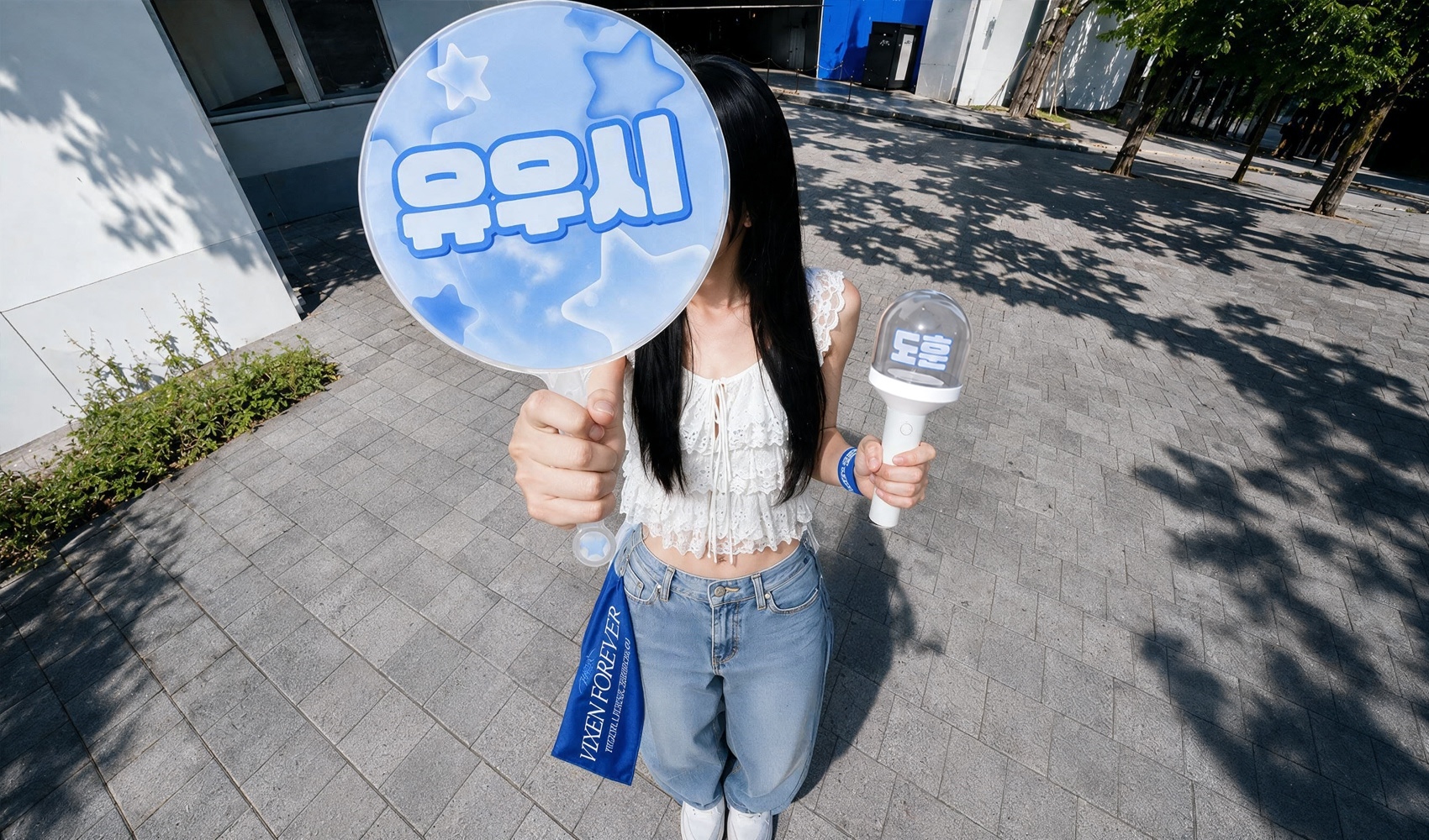 마플 콘서트 굿즈 추천 – 최애 이름을 넣은 대형 이미지 피켓과 LED 응원봉, 파란색 미니 슬로건을 든 여성이 야외 콘서트장 앞에서 촬영한 이미지